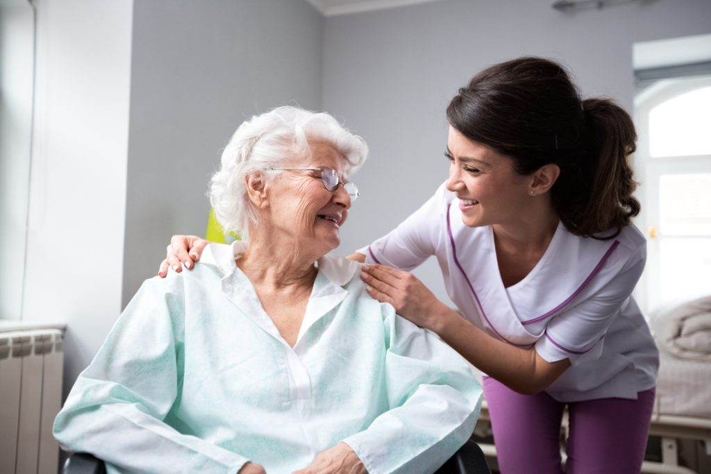 Nurse Smiling at Senior Woman on Chair and Holding her Shoulders
